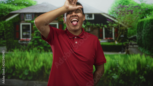 Фотография Hispanic man wearing red polo shirt showing tongue and making l sign on forehead at building entrance; playful mischief