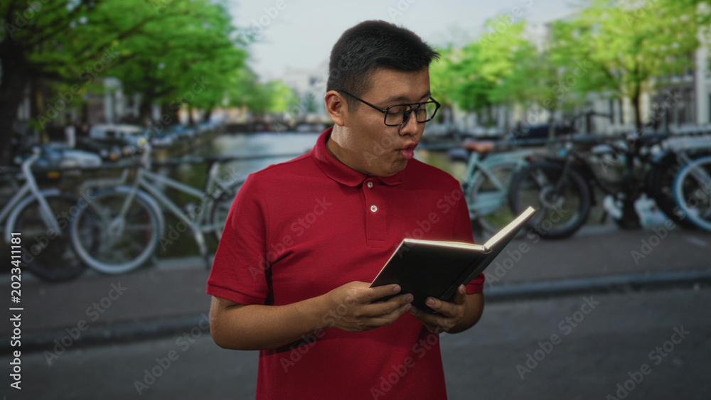 Obraz premium Man reading book on canal street beside bicycles and water, smiling while holding a notebook; urban leisure joy.