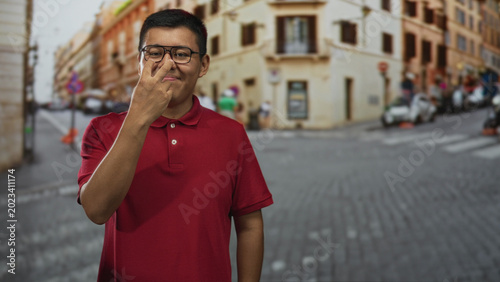 Quadro em tela Young man in red polo and glasses pinches his nose on a cobblestone street by old buildings and parked scooters, casual city scene; disgust annoyance