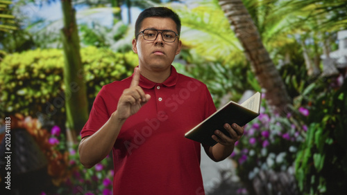 Photos Young hispanic man wearing glasses points index finger while holding an open book in a green park with palm trees and shrubs; quiet concentration