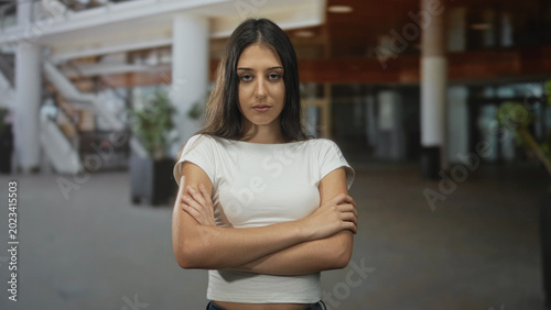 Photos Woman with crossed arms showing bare forearms and neutral expression in spacious building lobby with visible staircase and planters; defiance resilience