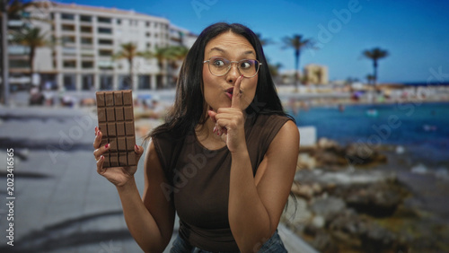Photos Woman holds chocolate bar up in hand by oceanfront building on a sunny boardwalk, wearing brown sleeveless top, glasses and jeans; curiosity