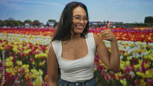 Fényképezés Woman wearing glasses and white tank top gives thumbs up showing bare shoulder in colorful tulip field, smiling at camera; joy