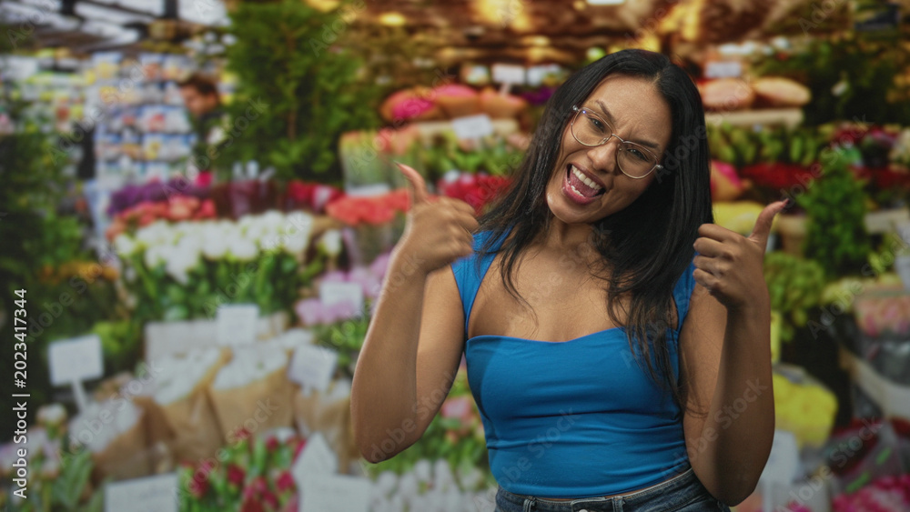 Fototapeta premium Woman in blue top showing thumbs up with visible thumbs and broad smile among bouquets at an indoor flower market building; joy confidence.