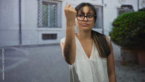 Tablou pe pânză Woman raising hand in a shrug gesture on a street in front of a building, glasses and white blouse visible; indifference casual attitude