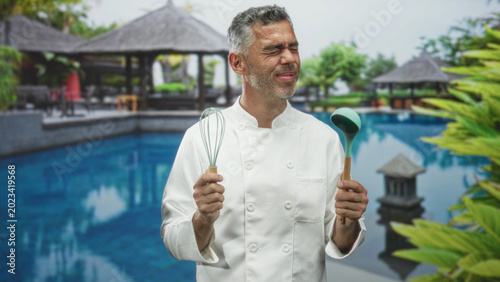 Valokuva Man chef holds whisk and ladle with hands, looks up laughing beside a hotel building pool; joyful celebration