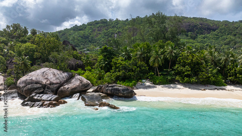 Foto Granite boulders situated by turquoise ocean waters, set against lush forested hills