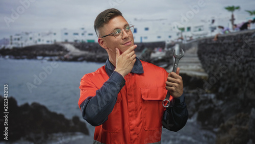 Fotografie Young mechanic man in orange coverall holding wrench, hand to chin gesture near coastal building and rocky pier; thoughtful pondering curiosity