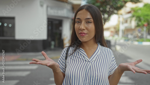 Foto Hispanic brunette woman shrugging with palms up on a city street crosswalk, wearing blue and white striped blouse and shoulder bag; indifference
