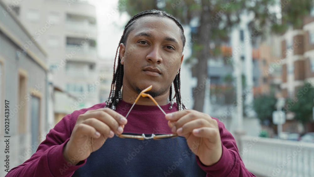 Fototapeta premium Man with braided beaded hair puts on sunglasses with both hands, maroon sweater visible, neutral face on street; confidence urban.