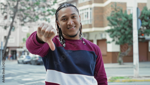 Fotografija Man giving thumb down gesture on street in front of apartment building while wearing burgundy and navy sweater and braided hair; disapproval casual