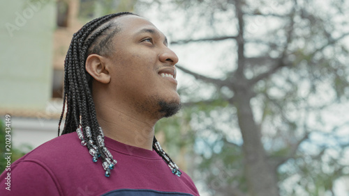 Konstfotografi Man young hispanic with braided hair and beaded ends looking up, smiling beside a building and tree; hopeful reflection