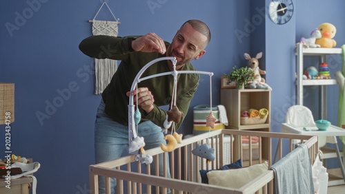 Konstfotografi Man leaning over wooden cradle adjusting plush mobile above mattress in studio baby room; bonding tenderness