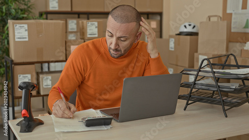 Man writing bills at a desk with laptop and cardboard packages, hand on head gesture in a parcel office building; small business stress.