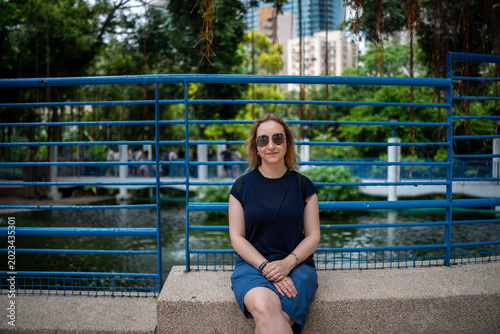 Woman sitting by a blue fence in a park with a pond and city buildings in background
