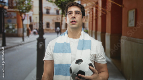 Foto Man holding soccer ball with blue and white striped scarf, standing on a narrow street near archways and a lamppost; anticipation fandom