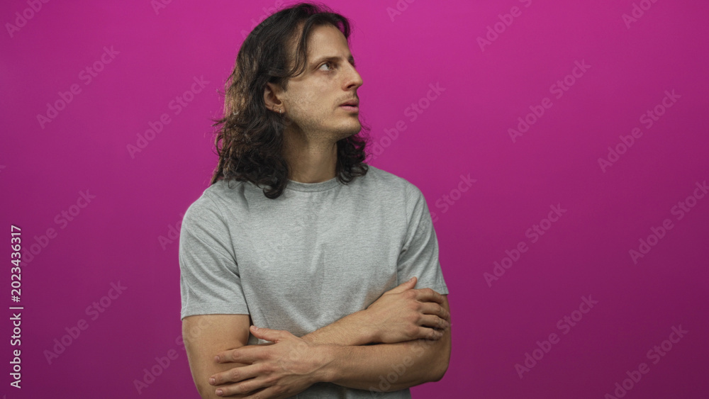 Obraz premium Young hispanic man with long hair and arms crossed in magenta studio wearing grey t shirt, looking sideways and contemplative; introspection.