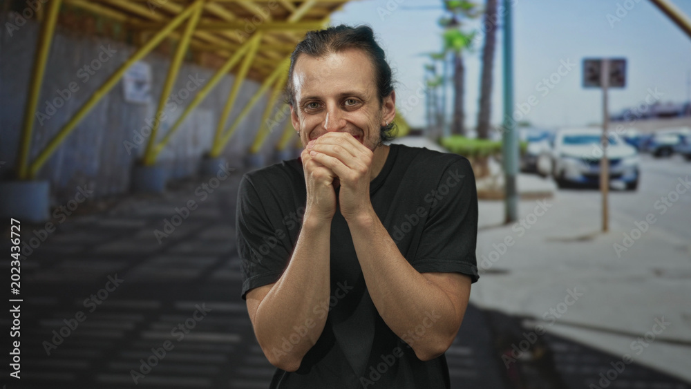 Fototapeta premium Man with hands over mouth smiling on street by parked cars and palm trees; nervousness anticipation.