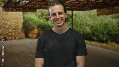 Konstfotografi Man smiling and seated on a wooden deck in a forest pavilion under a woven canopy; serenity ease openness