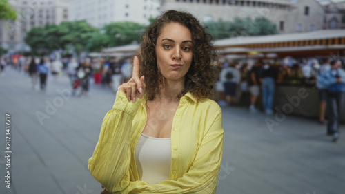 Canvas-taulu Woman points index finger upward in crowded city street market, wearing yellow shirt and white tank top with folded arm gesture; disapproval caution
