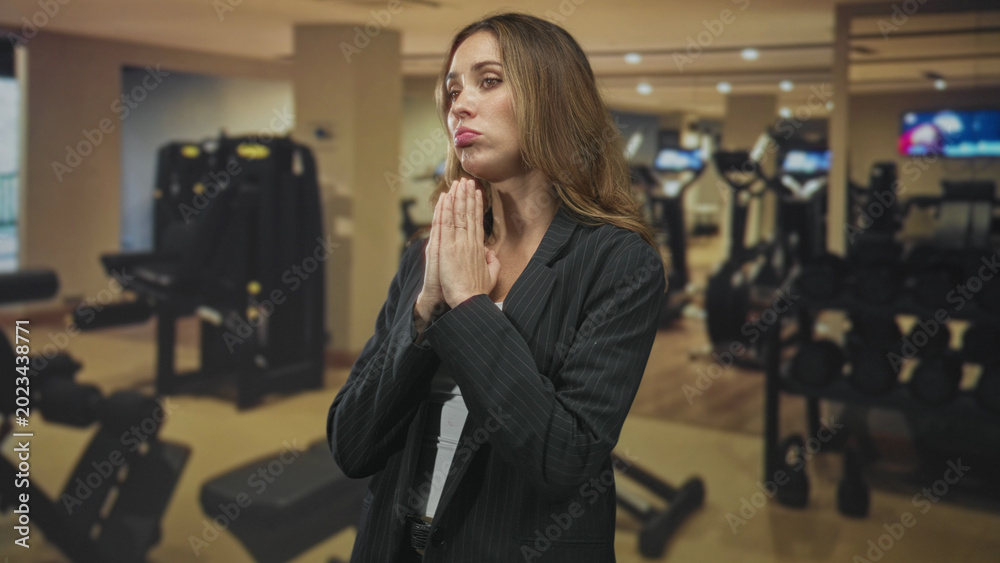 Fototapeta premium Woman with hands pressed in prayer gesture, pouting and wearing a blazer standing before dumbbells and a rack in a gym inside a building; pleading hope.