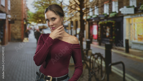 Quadro em tela Woman with bare chin touched by right hand in maroon off shoulder top on a cobbled street near bicycle rack and storefronts; confidence contemplation