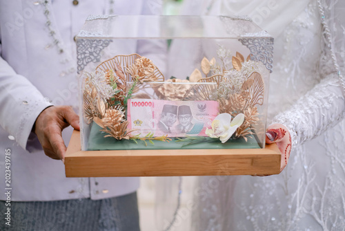 Принти на полотні A couple holding a decorative glass box containing Indonesian currency as a traditional wedding dowry during a formal marriage ceremony