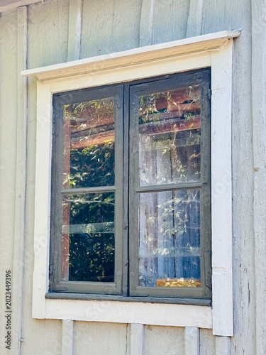Close-up of a traditional old wooden window set in a rustic farmhouse wall. Reflections of trees and sunlight create a nostalgic countryside atmosphere with Scandinavian rural charm.