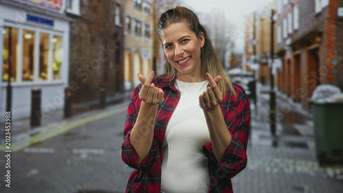 Foto Woman smiling makes finger heart with hands wearing red plaid shirt and white tee on a wet cobbled street with shopfronts and bins; playful