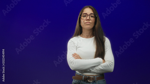 Photos Woman with glasses, arms crossed, long hair, wearing white top, jeans and brown belt against a deep purple studio backdrop; quiet determination