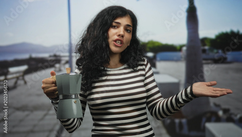 Quadro em tela Young woman holding a moka pot with one hand and speaking while gesturing with the other on a seaside street; casual