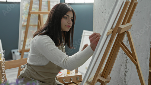Photos Young latin woman painter wearing apron leans forward and paints a canvas with a brush on a wooden easel in a studio; creativity concentration