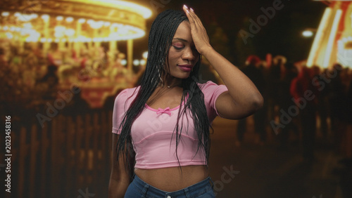 Valokuva Black woman with hand on forehead, pink crop top and visible midriff beside a lit carousel at a night street fair; playful nostalgia