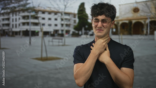 Bild auf Leinwand Young hispanic man clutching his throat with both hands on a city street plaza, grimacing and choking in pain; panic help needed