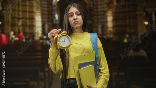 Quadro su tela Young female student in yellow sweater holds a yellow alarm clock and folders with a blue backpack, pouting while raising the clock in a building with pews; lateness anxiety