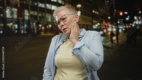 Fotografie Woman touching cheek with hand on a busy city street at night near lit shops wearing glasses and casual shirt; discomfort pain concern