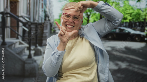 Fotografia Middle aged woman smiling and framing her face with both hands on a busy street by stoop steps and parked cars; playful joy