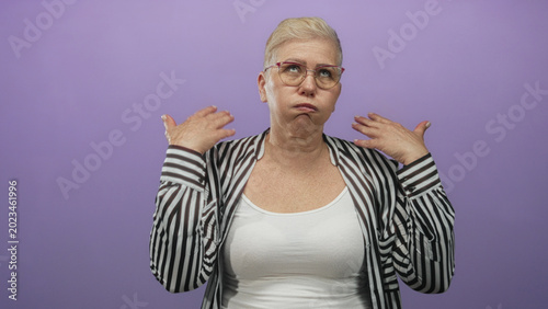 Canvas Print Woman with glasses short blonde hair wearing striped shirt and white tank top raising both hands to ears while pouting and looking up in purple studio; frustration