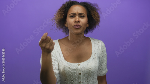 Photos Woman beckoning with hand and furrowed brow in purple studio wearing white knit top; confusion doubt