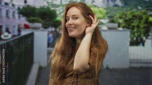 Photos Redheaded woman in brown dress holds hand to ear on busy street in daylight; curiosity awareness alertness