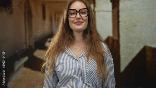 Foto Caucasian woman with black glasses adjusts frame while smiling softly in old town street under sunlight; confidence