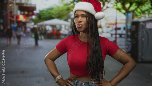 Photography Woman in red top and santa hat standing with hands on hips and pouting expression on a busy street; holiday annoyance