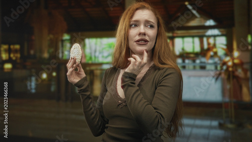 Fotografía Redhead woman holding pastry and touching chin in a warmly lit building interior, examining a cookie with a hesitant expression; hesitation decision