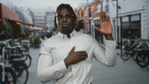 Fotografie Man wearing white shirt and glasses with hand on chest and right hand raised in street with bicycles and brick buildings; pledge solemn