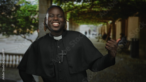 Quadro em tela Man priest gives thumbs up with bare hand in front of building colonnade, wearing clerical collar and cross necklace; joy faith outreach