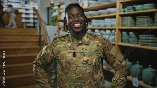 Fotografi Man soldier in camouflage uniform smiling with hands on hips and dogtag in home decor shop; pride duty