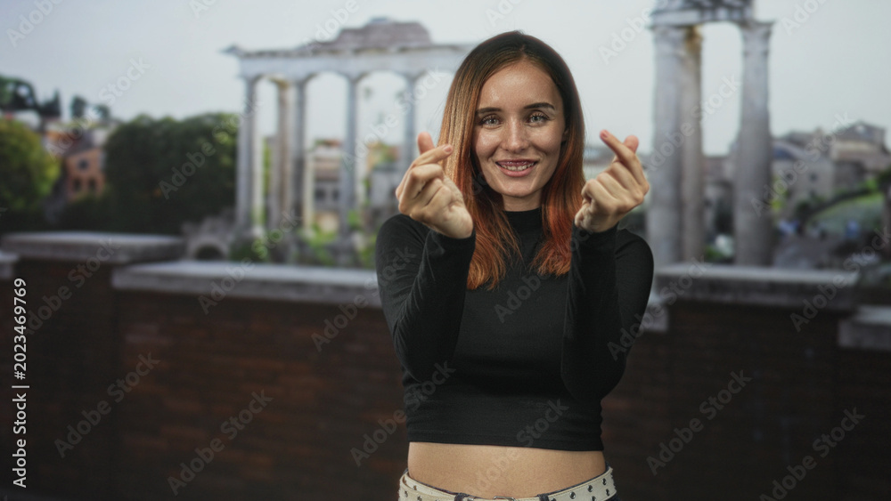 Obraz premium Young woman making twin finger heart gestures showing midriff at building columns and terrace; playful confidence.