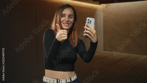 Fotografija Woman redhead smiling while holding smartphone and giving thumbs up in studio, visible midriff and studded belt; confidence connection
