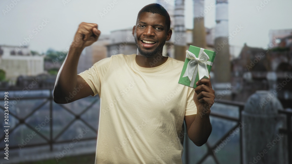 Fototapeta premium Man holding green gift with white bow and raised fist at building railing, smiling broadly; celebration victory.