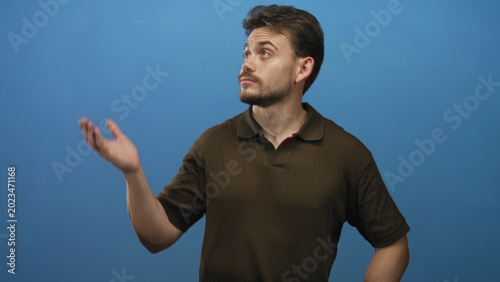 Quadro su tela Young hispanic man glancing sideways with hands on hips wearing brown polo shirt in blue studio; skepticism doubt contemplation